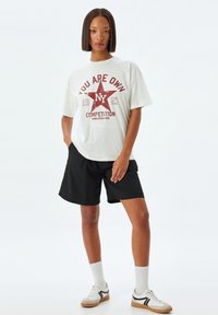 Camiseta blanca de gran tamaño con un estampado gráfico rojo que presenta una estrella y texto. Se lleva con pantalones cortos negros y calcetas blancas, combinados con zapatillas.