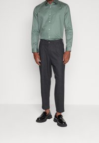 Herroutfit med en ljusgrön knäppskjorta och mörkgrå smala byxor. Skorna är glänsande svarta med en remdetalj.