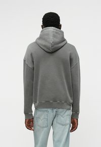 Γκρι, υπερμεγέθη φούτερ με μεγάλη κουκούλα, ριμπ μανσέτες και στρίφωμα, φτιαγμένο από μαλακό ύφασμα. Συνδυάζεται με ανοιχτό μπλε τζιν, δείχνοντας έναν χαλαρό σχεδιασμό.