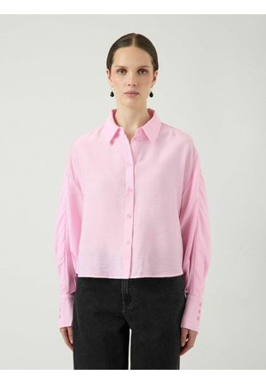 Kobieta ubrana w jasnoróżową koszulę na guziki z długim rękawem i dużymi mankietami oraz czarne spodnie, stojąca na jednolitym tle.