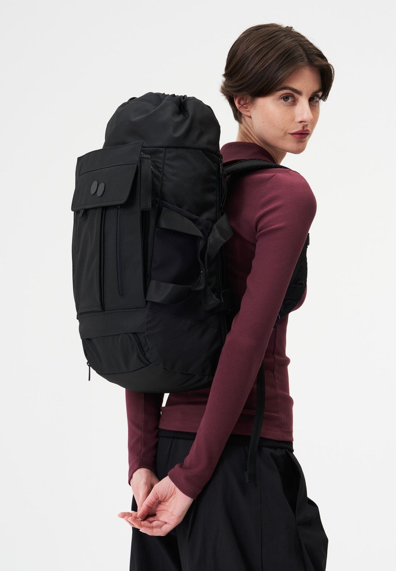 Femme portant un grand sac à dos noir à multiples poches, vêtue d'un chemisier à manches longues bordeaux et d'un pantalon noir, debout devant un fond blanc.