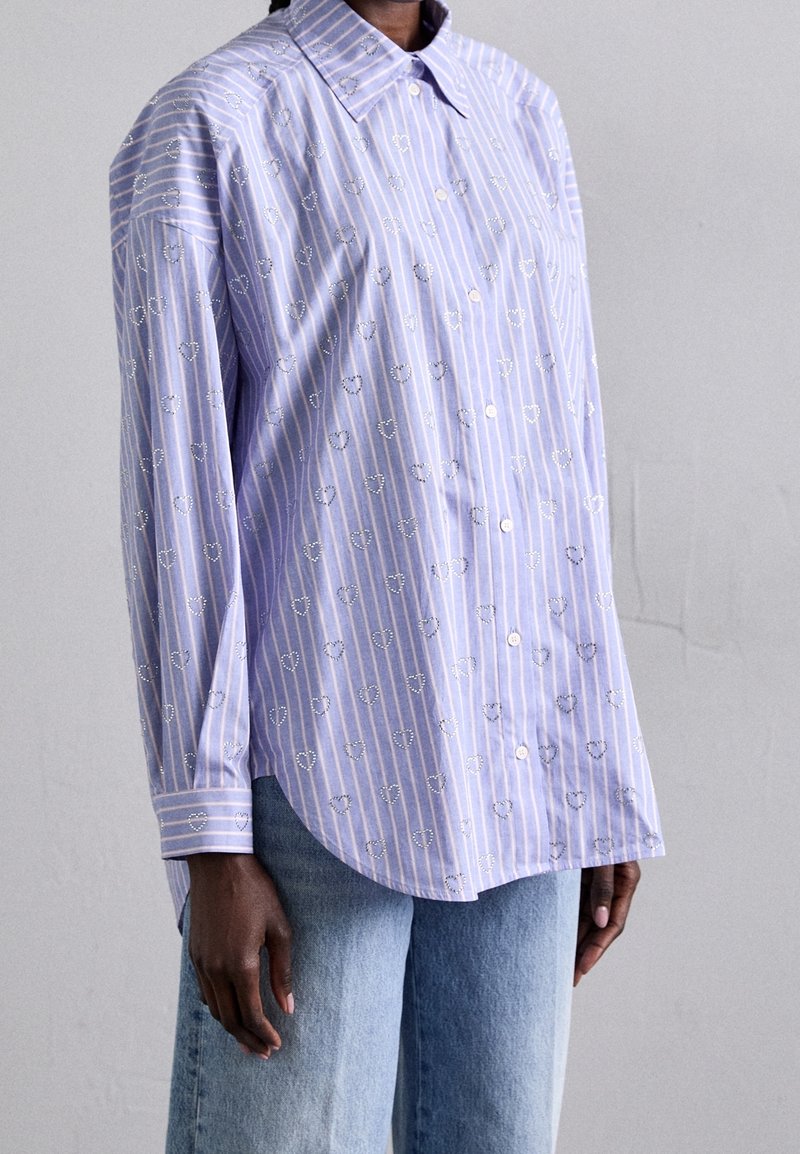 Femme portant une chemise à rayures bleu clair avec petits motifs en forme de cœur, associée à un jean bleu clair.