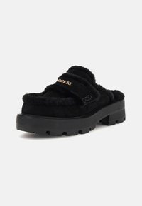 Čierne huňaté slip-on topánky s hrubou čiernou platformou, zdobené dvojitými pásmi a zlatým logom "GUESS" na vrchu.