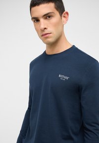 Tumši zils garām piedurknēm T-krekls no mīksta auduma, ar apaļu apkakli un mazu baltu logotipu kreisajā krūšu daļā.