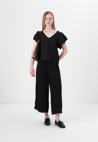 Black textured top with ruffled sleeves, paired with black wide-leg pants. Model carries a maroon shoulder bag, wearing black shoes.