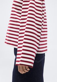 Κόκκινη και λευκή ριγέ μπλούζα με μακριά μανίκια και άνετη εφαρμογή, με στρογγυλεμένη κάτω ραφή και ορατές ραφές. Το ύφασμα φαίνεται μαλακό και υφασμένο.