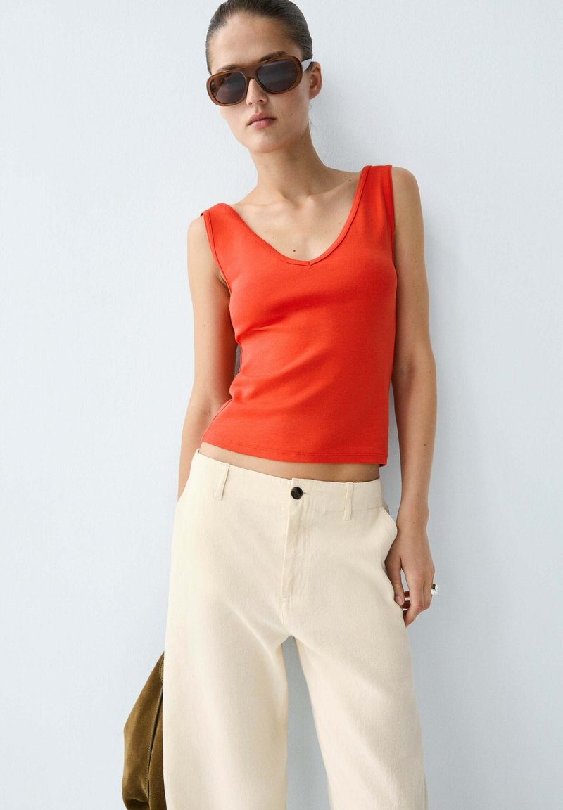 Woman wearing dark sunglasses, sleeveless red top, and high-waisted loose beige trousers, standing against a white wall.
