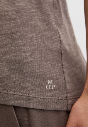 Gros plan sur une personne portant une chemise en maille marron avec le logo "M O P" près de l'ourlet et un pantalon marron assorti.