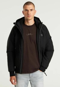 Črna zip-up jakna s kapuco, teksturirana tkanina. Ima stranske žepe in logotip. Nosi se čez rjav srajco in svetle kavbojke.