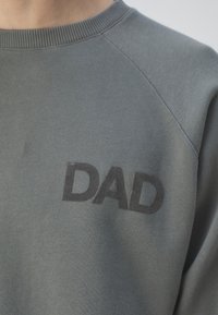 Detailní záběr šedé mikiny s kulatým výstřihem, na které jsou černými písmeny napsána slova "DAD" na hrudi.