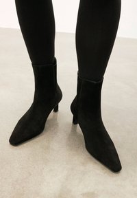 Melnas zamšādas potītes zābakus ar smailiem purngaliem, īsiem papēžiem un sānu rāvējslēdzējiem. Zābakiem ir gluda tekstūra un eleganta dizaina.