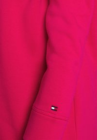 Vêtement à manches longues rose vif avec un petit logo rectangulaire rouge, blanc et bleu marine brodé près du poignet.