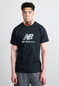 T-shirt in cotone nero con logo "New Balance" argentato sul davanti. Il design include maniche corte e collo rotondo.