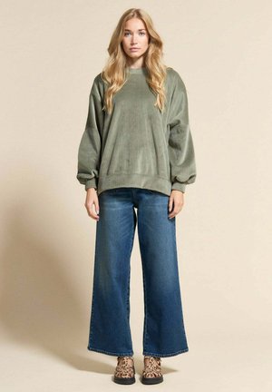 Femme aux longs cheveux blonds portant un sweat-shirt vert oversize, un jean large bleu et des chaussures à imprimé léopard, debout devant un fond beige.