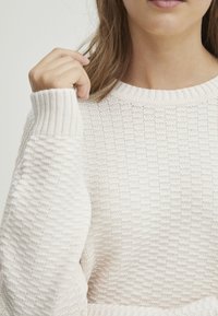Helles, cremigen Strickpullover mit strukturiertem Muster, gerippten Bündchen und rundem Ausschnitt. Nahaufnahme des Schulter- und Handdetails.