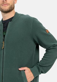 Grüne strukturierte Zip-Cardigan mit gerippten Bündchen, ausgestattet mit einem Lederlogo-Patch am Ärmel und seitlichen Taschen.