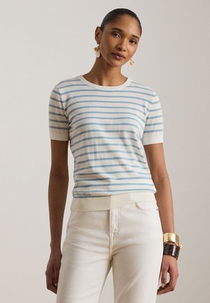 Naine kannab hele sinise ja valge triibulist lühikese varrukaga kampsunit, valgeid pükse ning vasakul randmel kuld- ja kilpkonnamustriga käevõrusid.