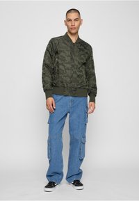Veste bomber camouflage vert olive avec poignets et ourlet côtelés, associée à un pantalon cargo ample bleu clair avec poches latérales et des baskets noires.