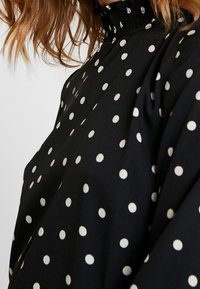 Blusa nera con pois bianchi, caratterizzata da un alto colletto arricciato e maniche ampie e fluenti, realizzata in un tessuto morbido con una superficie liscia.