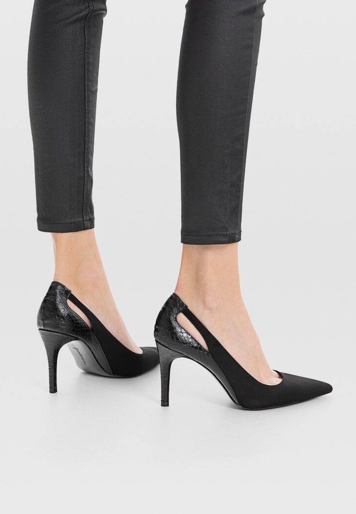 Jambes portant un pantalon skinny noir et des chaussures à talons hauts noires avec des bouts pointus et des panneaux arrière texturés sur un fond blanc.