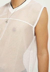 Λευκό αμάνικο τοπ από δίχτυ με μαύρες λεπτομέρειες, φορεμένο από άτομο με μαυρισμένο δέρμα και εμφανές ασημένιο μενταγιόν στο λαιμό.