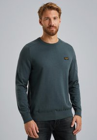 Dunkelgrüner Pullover aus weichem Stoff, Rundhalsausschnitt, gerippte Bündchen und Saum, mit einem kleinen Markenpatch auf der Brust. Getragen mit dunklen Jeans.