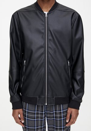 Homme portant une veste aviateur zippée en similicuir noir et un pantalon à carreaux gris avec des rayures bleues et blanches, debout devant un fond blanc.