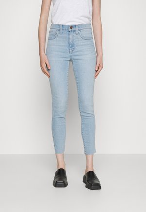 Personne portant un jean skinny taille haute bleu clair, un t-shirt blanc et des chaussures noires slip-on, debout devant un fond uni et clair.