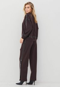 Moteris tamsiai rudos spalvos sportinėje aprangoje su blizgiomis juodomis detalėmis ir juodais aukštakulniais batais, stovi ir žiūri per petį prie baltos sienos.