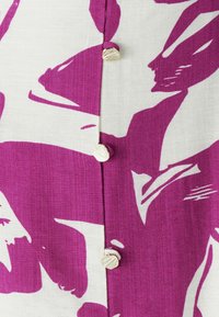 Blouse à manches courtes en tissu blanc avec un motif abstrait violet audacieux, fermée par des boutons uniques en forme de coquillage sur le devant.