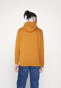 Sweatshirt à capuche orange avec des manches longues, texture lisse et poignets côtelés, associé à un pantalon bleu. Conception simple et décontractée.