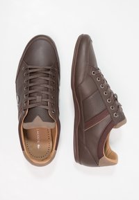 Bruna lädersneakers med texturerade sidor, snörningssystem och vadderad krage; har kontrasterande bruna accenter och en slät yta.