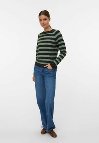 Maglione a righe in verde scuro e nero, scollo a cerchio, maniche lunghe, abbinato a jeans a gamba larga blu e scarpe marroni.