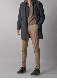 Cappotto blu scuro con chiusure a bottone, indossato sopra una giacca beige con zip e pantaloni marroni. Completato da sneakers verdi, creando un look informale.
