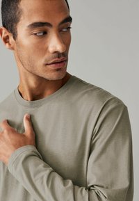 Langsleeve shirt in gedempt olijf groen katoen. Ronde halslijn, relaxte pasvorm, gladde textuur en zichtbare stiksel-details op de schouders.