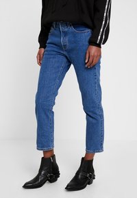 Vaqueros de mezclilla azul con cintura alta, diseño de pierna recta, dobladillo con vuelta y estilo clásico de cinco bolsillos, combinados con botas negras hasta el tobillo.