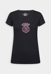 Camiseta de algodón negra con cuello redondo, que presenta un logo bordado en rosa de "GUESS" y un diseño de escudo en la parte delantera. Mangas cortas, forma ajustada.