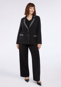 Costum negru format dintr-un sacou croit cu rever texturat și pantaloni cu picioare largi asortați. Accente texturate pe rever și buzunare.