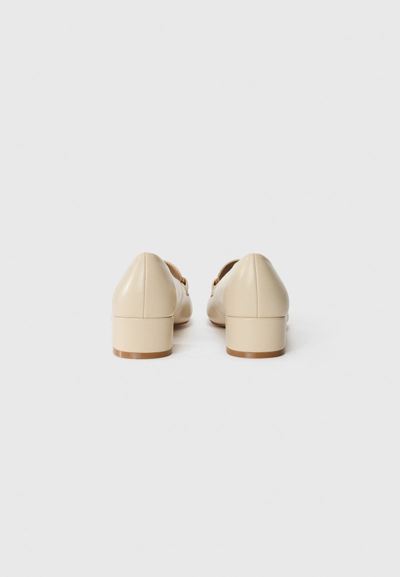 Chaussures à talons blocs en cuir beige présentant un bout pointu et une finition glossy subtile. Agrémentées de quincaillerie dorée sur les côtés.
