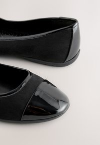 Paar schwarze Damenschuhe mit flacher Sohle, Wildlederseiten und glänzenden Lacklederkappen an Zehen und Fersen auf einer hellen Oberfläche.
