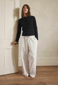 Woman standing barefoot in white polka dot pants and black buttoned shirt, leaning on partially open white door in neutral-toned room.