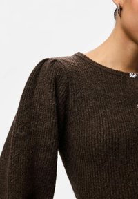 Brūns teksturēts džemperis ar ribotām detaļām, apaļu kakla izgriezumu un pūkainām piedurknēm. Iekļauj dekoratīvos pogu elementus priekšpusē.