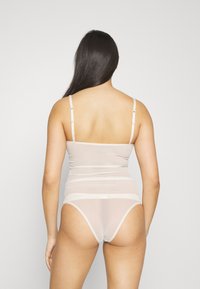 Bodysuit transparent beige clair avec bretelles ajustables, texture lisse et rayures horizontales au niveau de la taille. Dos en thong à taille haute.