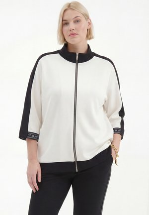 Vrouw met kort blond haar, die een zwart-witte jas met rits en driekwart mouwen draagt, zwarte broek en een gouden armband.