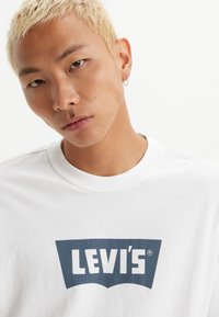 Balta medvilnės marškinėliai su mėlynu Levi's logo ant krūtinės, su apvaliu iškirpimu ir trumpomis rankovėmis. Lygus, vienspalvis paviršius.