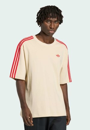 Giovane uomo con dreadlocks corti che indossa una t-shirt beige Adidas con strisce rosse sulle maniche e il logo sul petto, in piedi davanti a uno sfondo semplice.