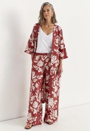 Kvinde iført en rust-rød blomsterkimono og vidde bukser med en hvid camisole og guldøreringe, stående mod en hvid baggrund.