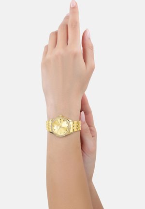 Goldene Armbanduhr mit rundem Ziffernblatt, goldfarbigem Metallarmband und einer mit Kristallen verzierten Lünette. Sie verfügt über ein minimalistisches Ziffernblatt mit römischen Ziffern.