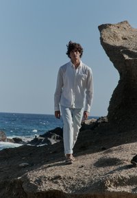 Chemise blanche boutonnée et jean bleu clair portés par une personne marchant sur un terrain rocheux au bord de l'océan, avec des vagues en arrière-plan.