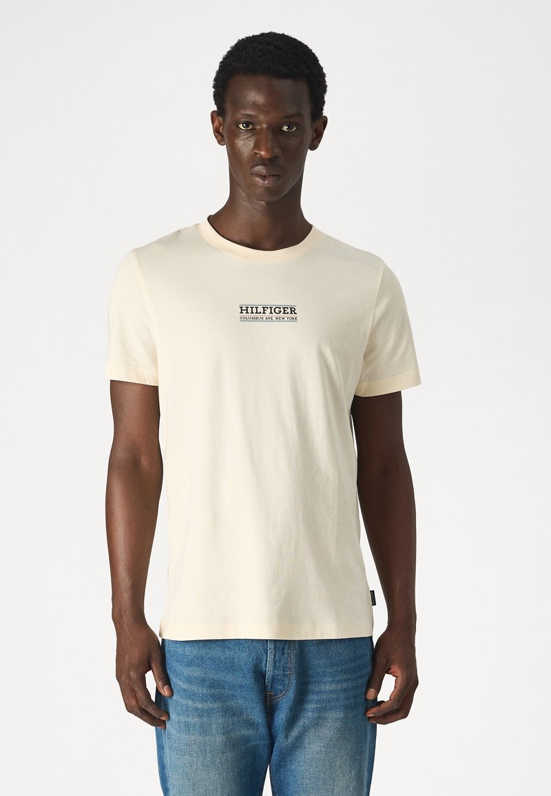 T-shirt in cotone crema con maniche corte, scollatura rotonda e un logo centrale "HILFIGER COLUMBUS AVE NEW YORK" in caratteri scuri.